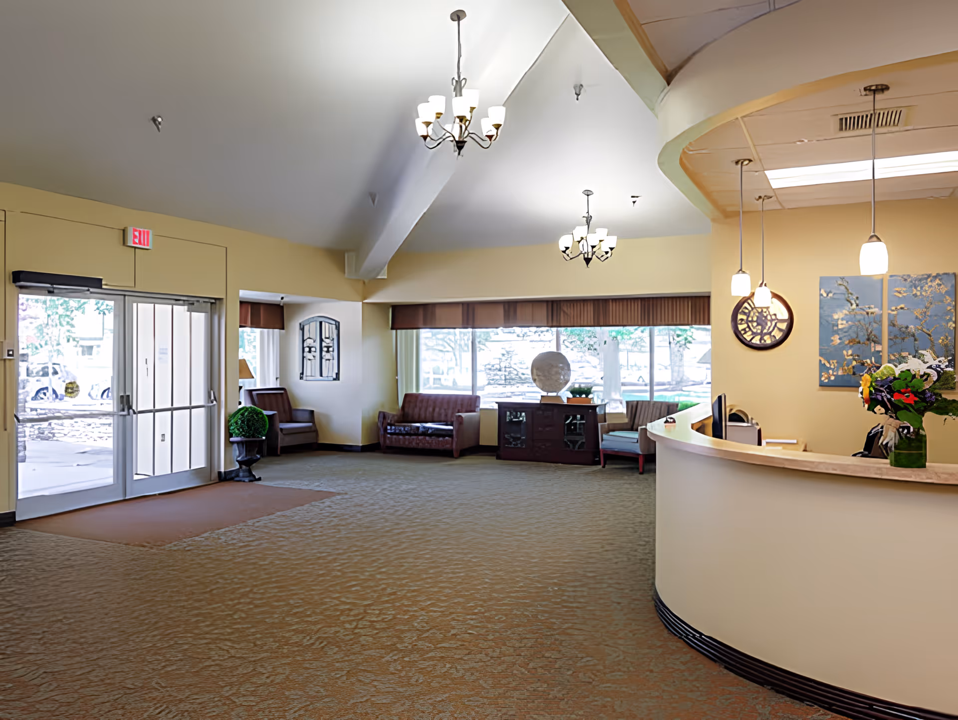 A spacious senior living facility lobby with beige walls and carpeted floor. The area features a curved reception desk with a vase of flowers, several chandeliers hanging from the ceiling, large windows letting in natural light, and seating areas with sofas and chairs near the windows. An exit door is visible on the left side.