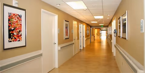 A long, well-lit hallway in a healthcare facility with beige walls and light-colored flooring. The hallway features several closed white doors on both sides, handrails along the walls, framed artwork hanging on the walls, and ceiling lights. The corridor extends into the distance with an exit sign visible at the far end.