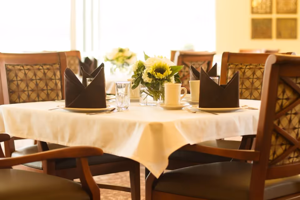 A dining table set with a white tablecloth, black folded napkins, clear drinking glasses, white coffee cups, and a centerpiece of yellow and white flowers in a glass vase. The table is surrounded by wooden chairs with patterned cushions, and more tables and chairs are visible in the softly lit background.