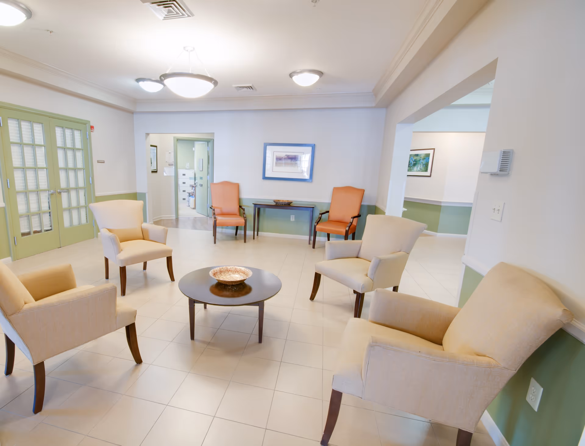 Bright seating area in a senior living facility with beige armchairs arranged around a round coffee table in a tiled lobby.