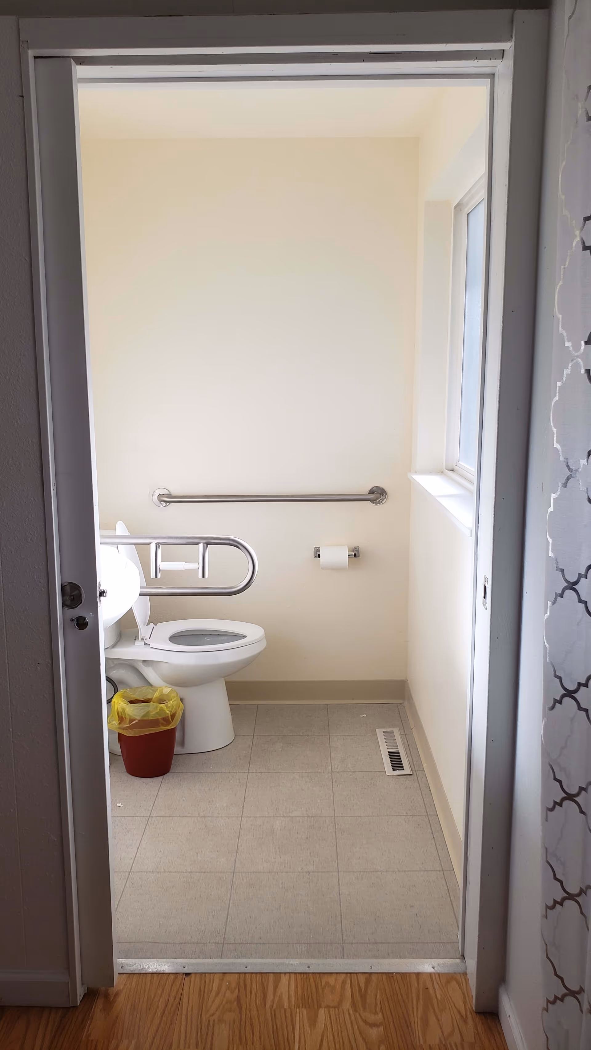 View into a small bathroom with a white toilet, a metal grab bar on the wall and another grab bar attached to the toilet. There is a red trash bin with a yellow liner next to the toilet. The floor is tiled and there is a window on the right wall.