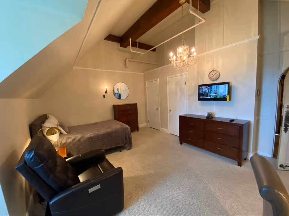 A cozy bedroom with a single bed covered in a gray blanket, a black recliner chair, a wooden dresser with a round mirror above it, and another larger wooden dresser with a wall-mounted TV and a clock above it. The room features beige walls, a chandelier hanging from the ceiling, and carpeted flooring.
