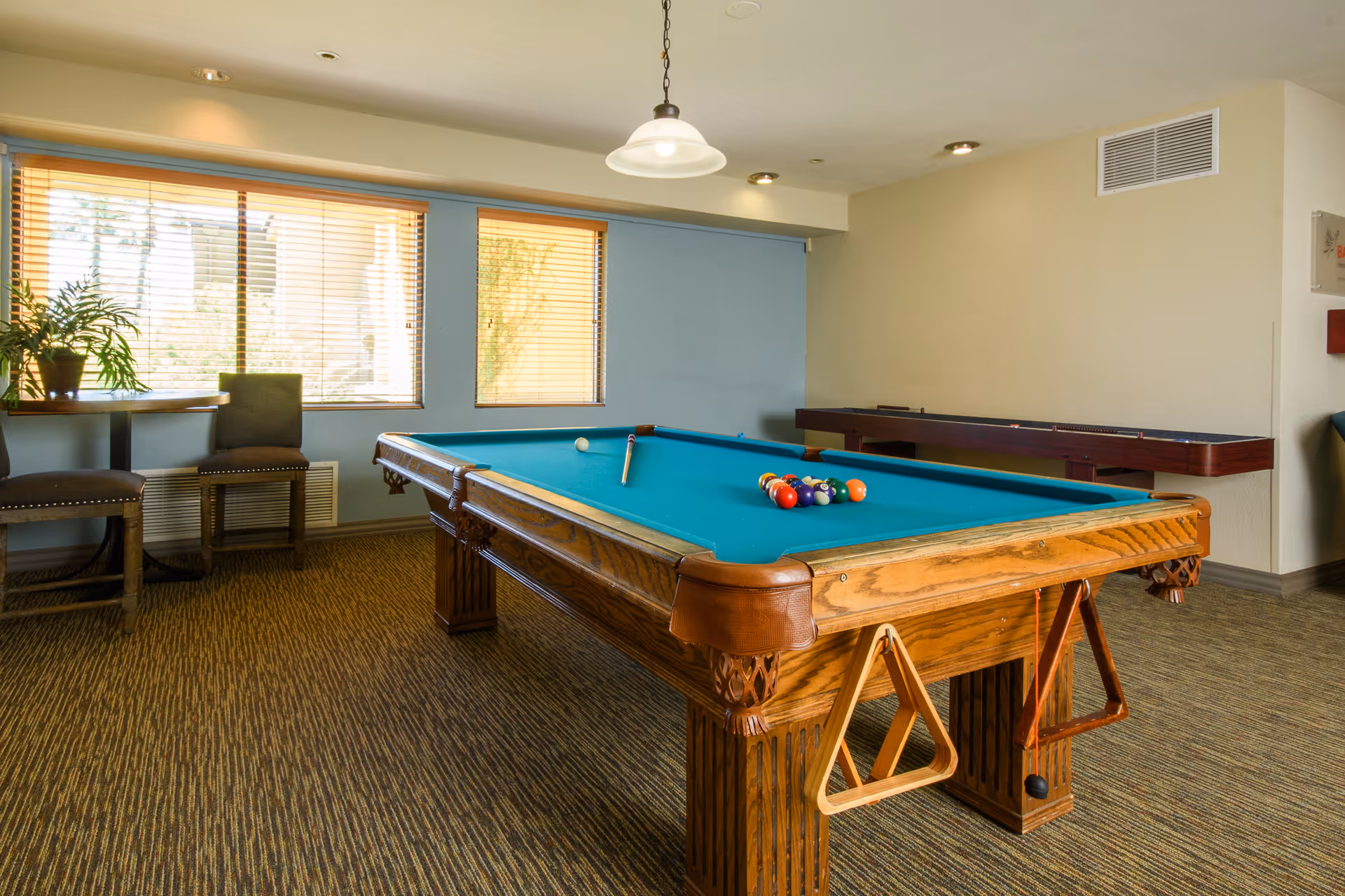 A bright recreation room interior with a wooden billiards table covered in teal felt, a cue and racked balls, seating by windows and a shuffleboard table along the wall.
