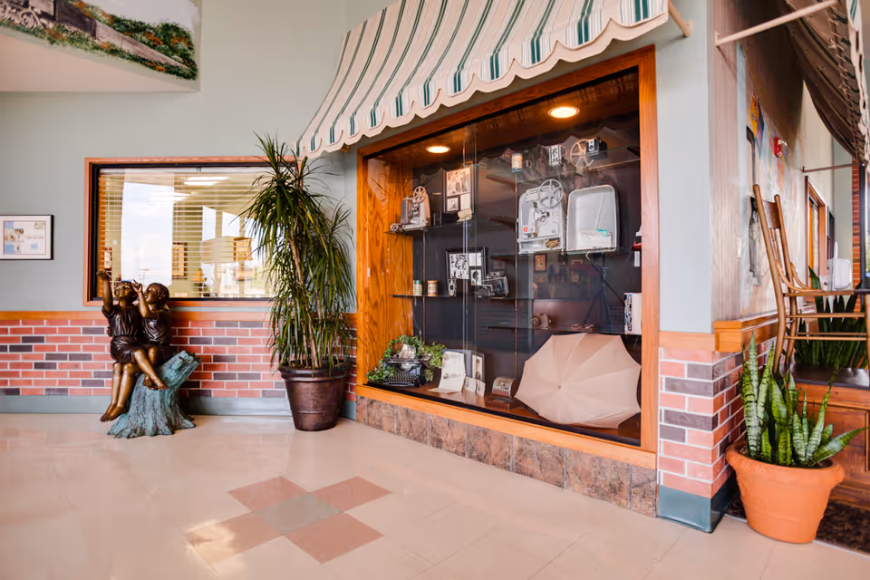 Interior view of a facility hallway featuring a display case with vintage film equipment and memorabilia, a bronze statue of two children sitting on a tree stump, potted plants, and a window with blinds. The walls have a brick lower half and light-colored upper half, with a striped awning above the display case.
