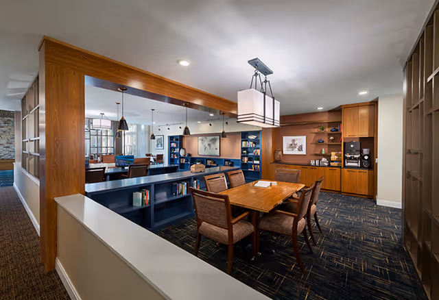 A spacious and well-lit common area in a senior living facility featuring a wooden table with six chairs, built-in bookshelves, and a coffee station with cabinets. The room has modern lighting fixtures and a carpeted floor with a patterned design. In the background, there are additional seating areas and large windows allowing natural light to enter.