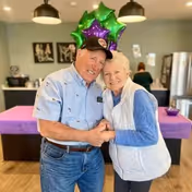 An elderly man and woman smiling and holding hands in a warmly lit room decorated with green and purple star-shaped balloons. The man is wearing a light blue shirt and a black cap, and the woman is wearing a white vest over a blue long-sleeve shirt. In the background, there is a kitchen area with a refrigerator and a person standing near the counter.