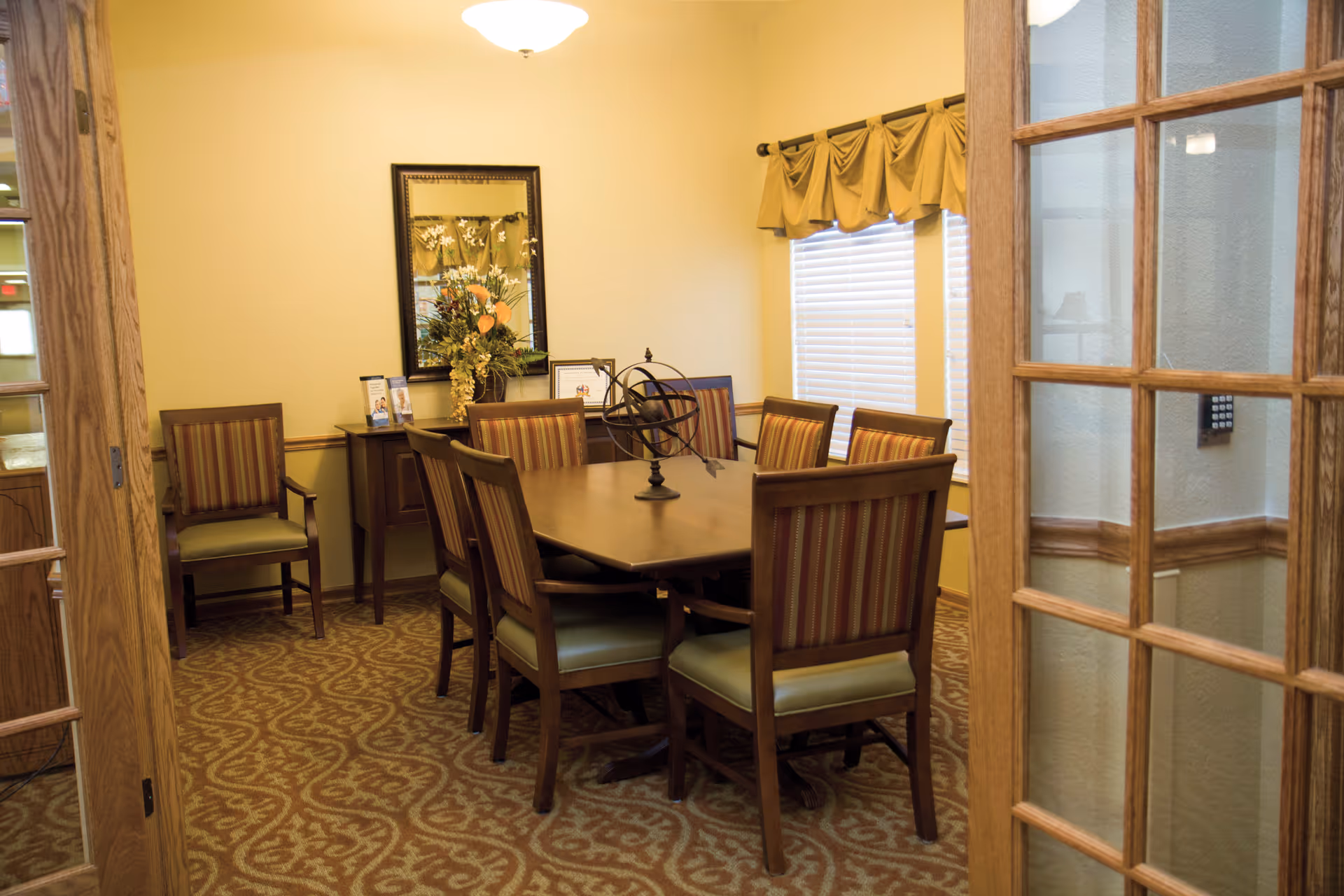 A small conference or meeting room with a rectangular wooden table surrounded by eight wooden chairs with striped upholstery. The room has beige walls, a patterned carpet, a window with blinds and a valance, a wall mirror above a sideboard with decorative items including a flower arrangement, and a ceiling light fixture. The room is viewed through partially open wooden French doors with glass panes.