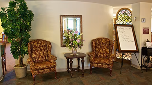 Two upholstered floral armchairs flank a small table with a flower arrangement beneath a mirror in a facility lobby, with a potted plant and an easel sign nearby.