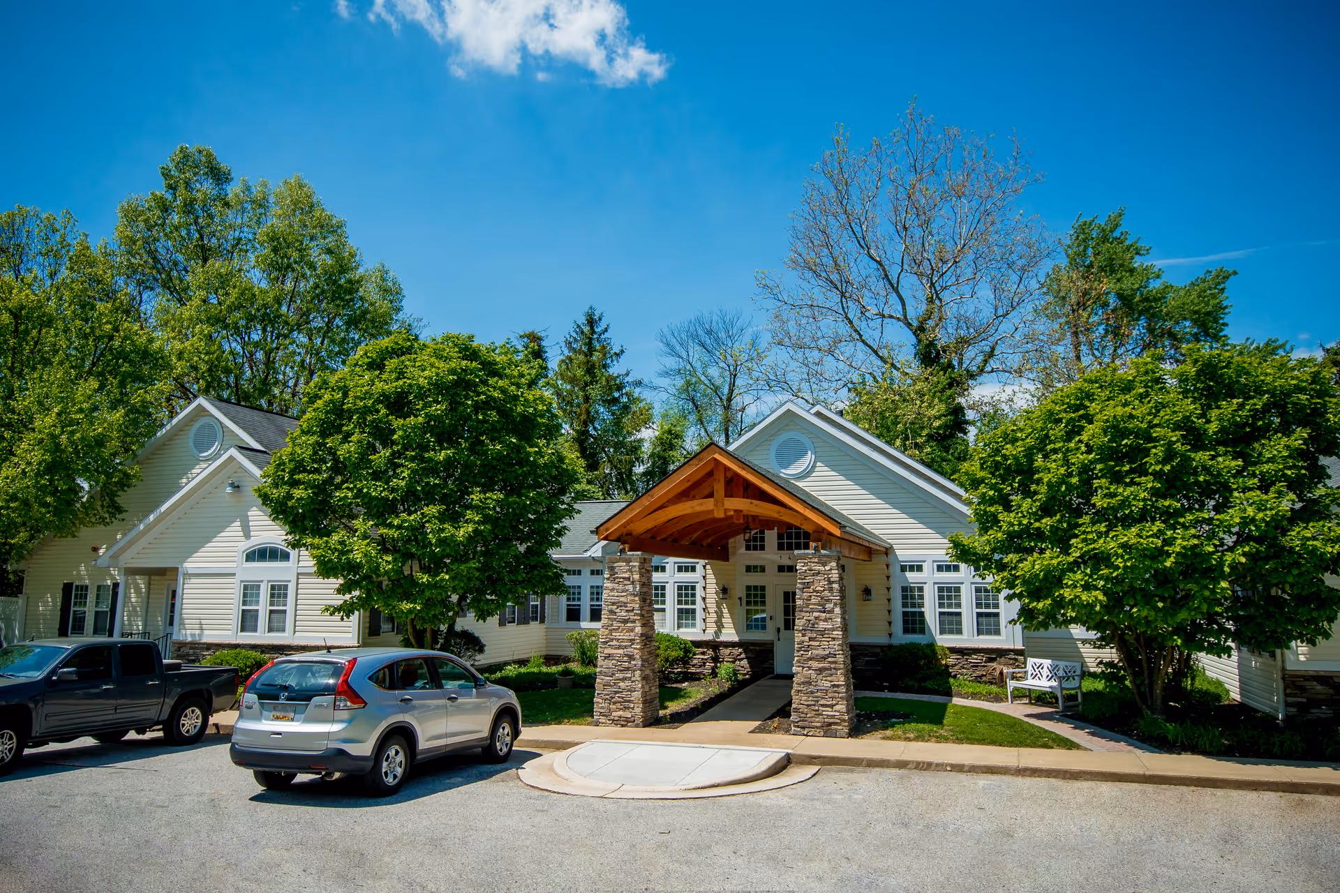 Exterior view of a single-story building with beige siding and a peaked roof, featuring a covered entrance supported by stone pillars. There are two cars parked in front, surrounded by green trees and a clear blue sky.