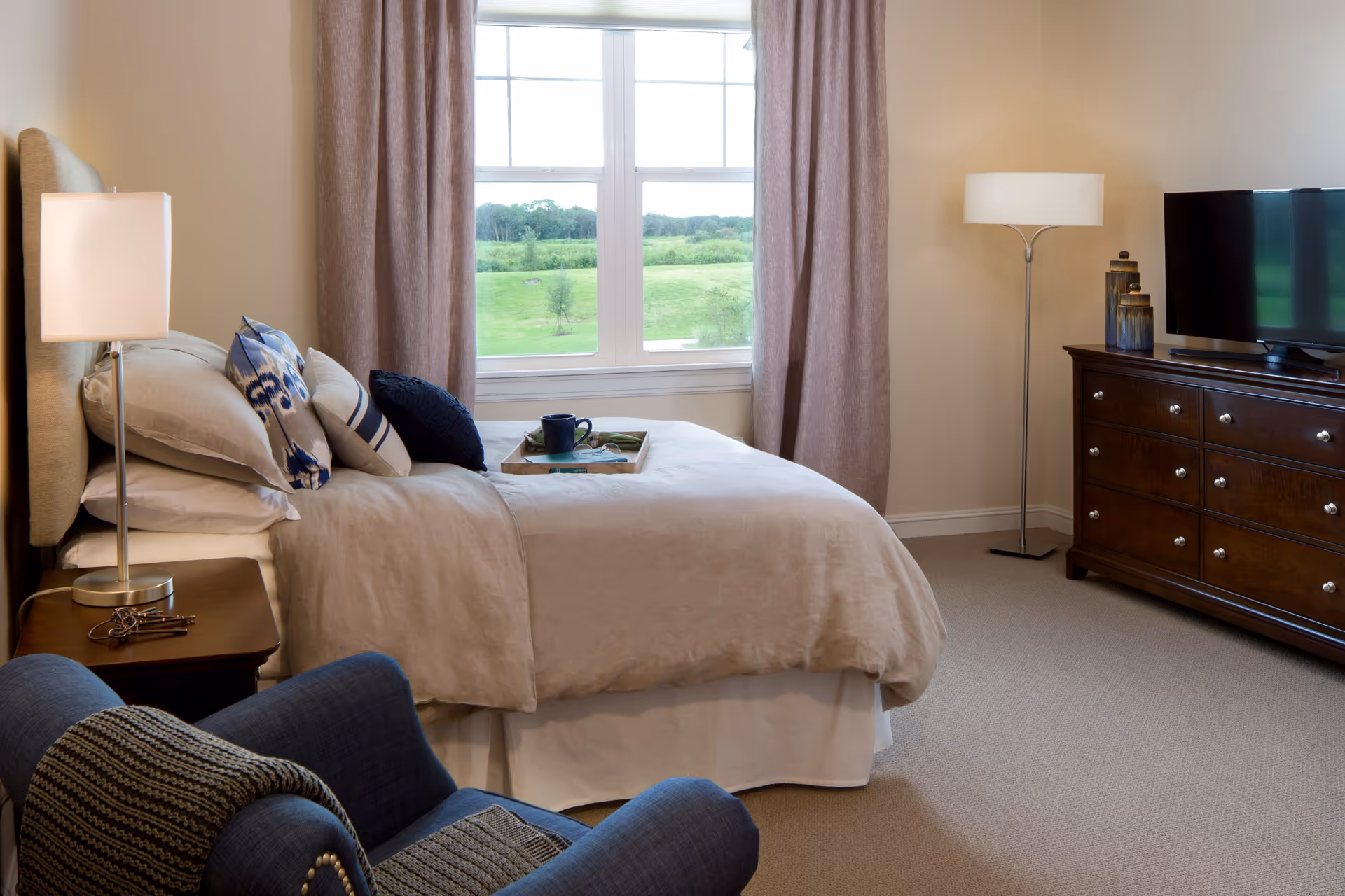 A cozy bedroom with a neatly made bed featuring beige bedding and decorative pillows. A wooden nightstand with a lamp and keys sits beside the bed. A blue armchair with a knitted throw is in the foreground. A large window with beige curtains shows a green outdoor landscape. Across from the bed is a dark wooden dresser with a flat-screen TV and decorative items. A floor lamp stands in the corner.