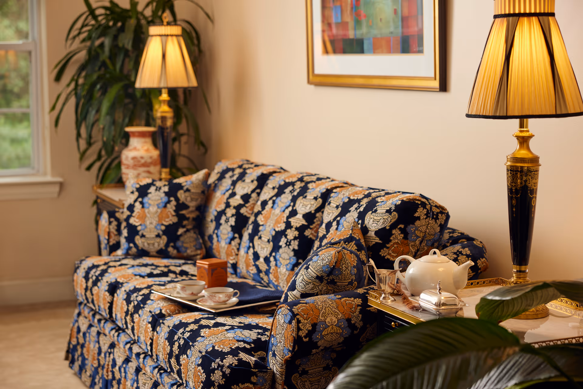 A cozy living room area with a patterned blue and gold sofa adorned with matching cushions. A tray with a teapot and teacups is placed on the sofa. There are two elegant table lamps with pleated shades on either side of the sofa, and a large leafy plant is visible in the background near a window. A framed colorful abstract painting hangs on the wall above the sofa.