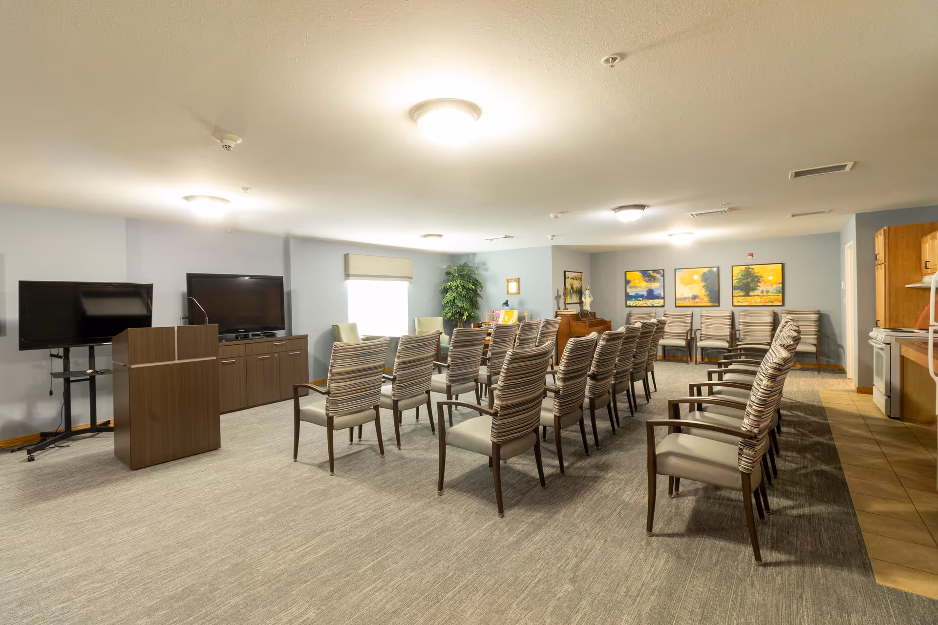 A well-lit room with rows of striped cushioned chairs arranged facing a podium and two large flat-screen TVs. The room has light-colored walls, carpeted flooring, and a window with a shade. There are framed pictures on the far wall and a small kitchen area visible on the right side.
