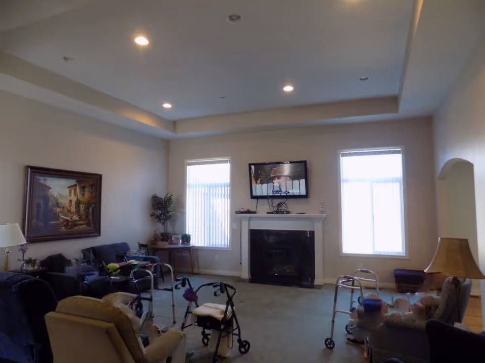 A senior living common room with armchairs, walkers, a fireplace, and a wall-mounted TV between two windows.