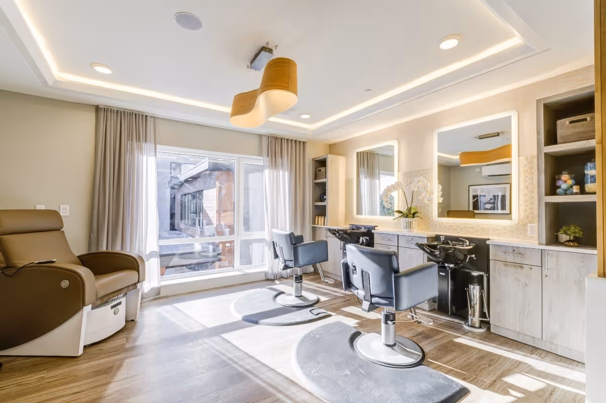 Bright and modern salon area with two black styling chairs in front of large illuminated mirrors, a comfortable brown recliner chair near a large window with sheer curtains, wooden flooring, and built-in cabinetry with shelves holding decorative items and hair products.