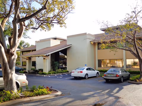 Exterior view of Plaza of Hallandale Beach Assisted Living facility showing a beige two-story building with large windows, a covered entrance, several parked cars, and trees with some greenery around the parking lot.