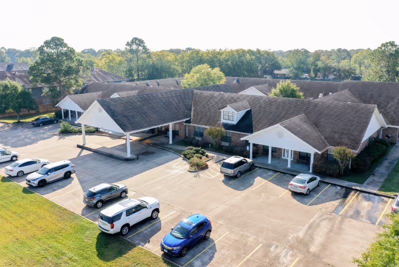Aerial view of Cambridge Square Assisted Living facility showing a single-story building with a covered entrance, surrounded by a parking lot with several parked cars and green trees in the background.