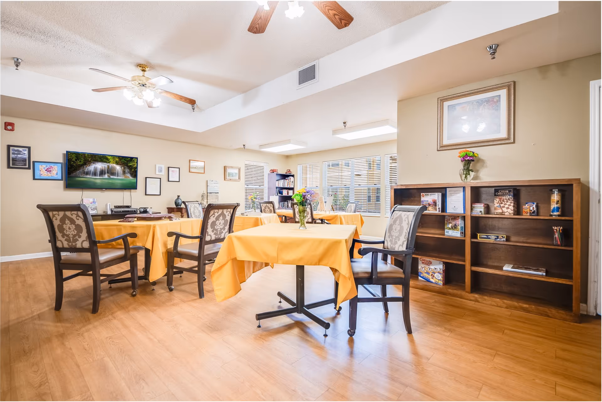 A bright and cozy common room with several tables covered in yellow tablecloths, each surrounded by cushioned chairs. The room features wooden flooring, a ceiling fan with lights, framed pictures on the walls, a flat-screen TV displaying a waterfall scene, and a bookshelf filled with games and books. Large windows allow natural light to fill the space.