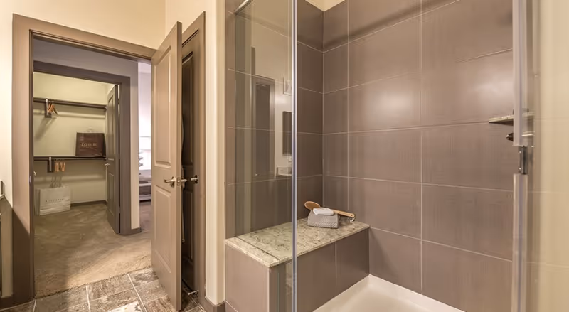 Modern tiled bathroom with a glass-enclosed shower and built-in bench, an open door revealing a walk-in closet and a glimpse of the adjoining bedroom.