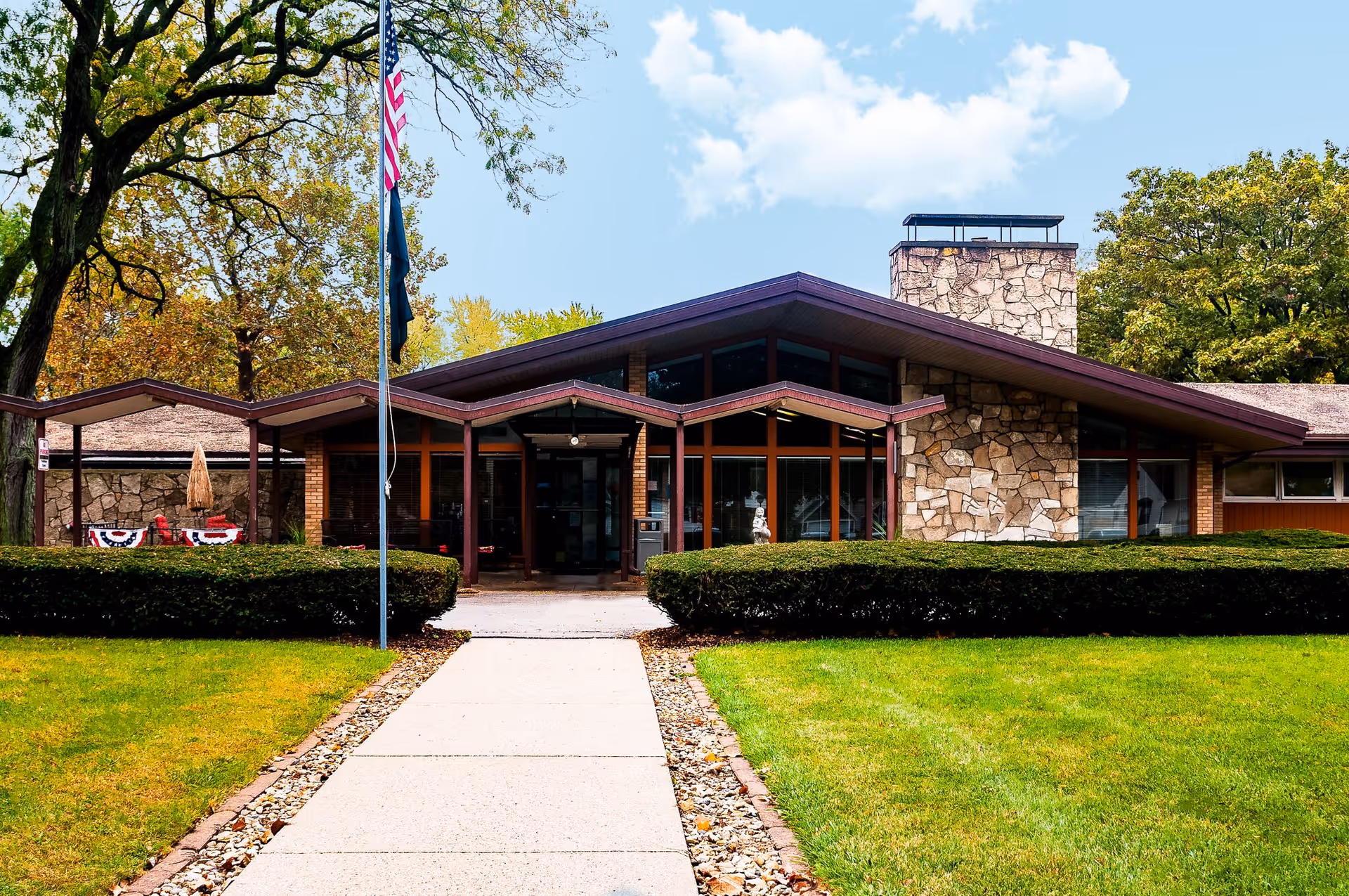 Front entrance of a low-slung stone-and-wood building with a walkway, flagpole, hedges, and lawn.