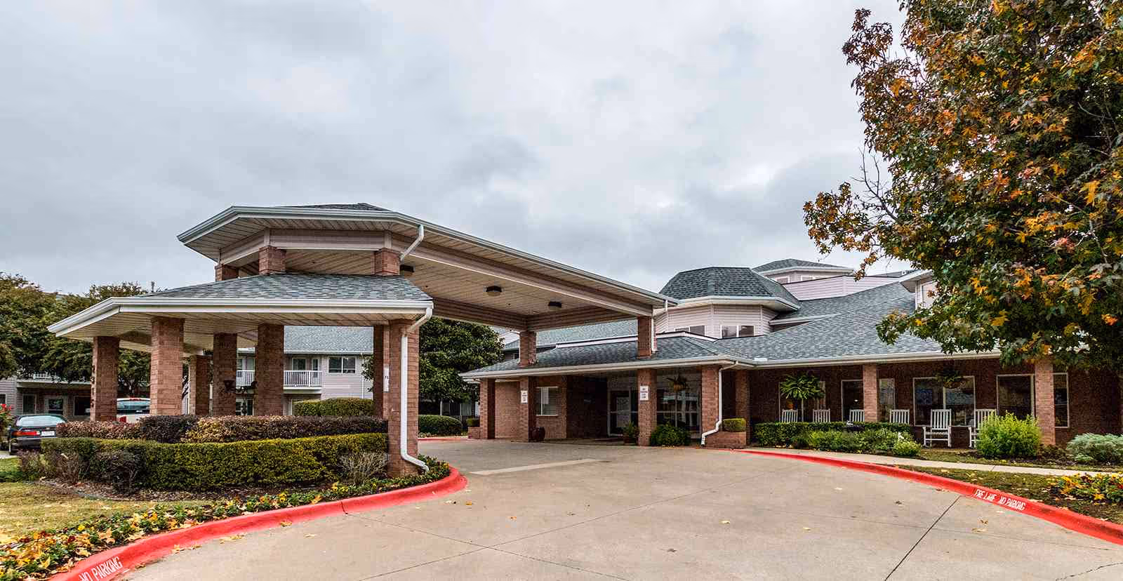 Covered entrance canopy and driveway in front of a brick senior living building with landscaping and rocking chairs on the porch.