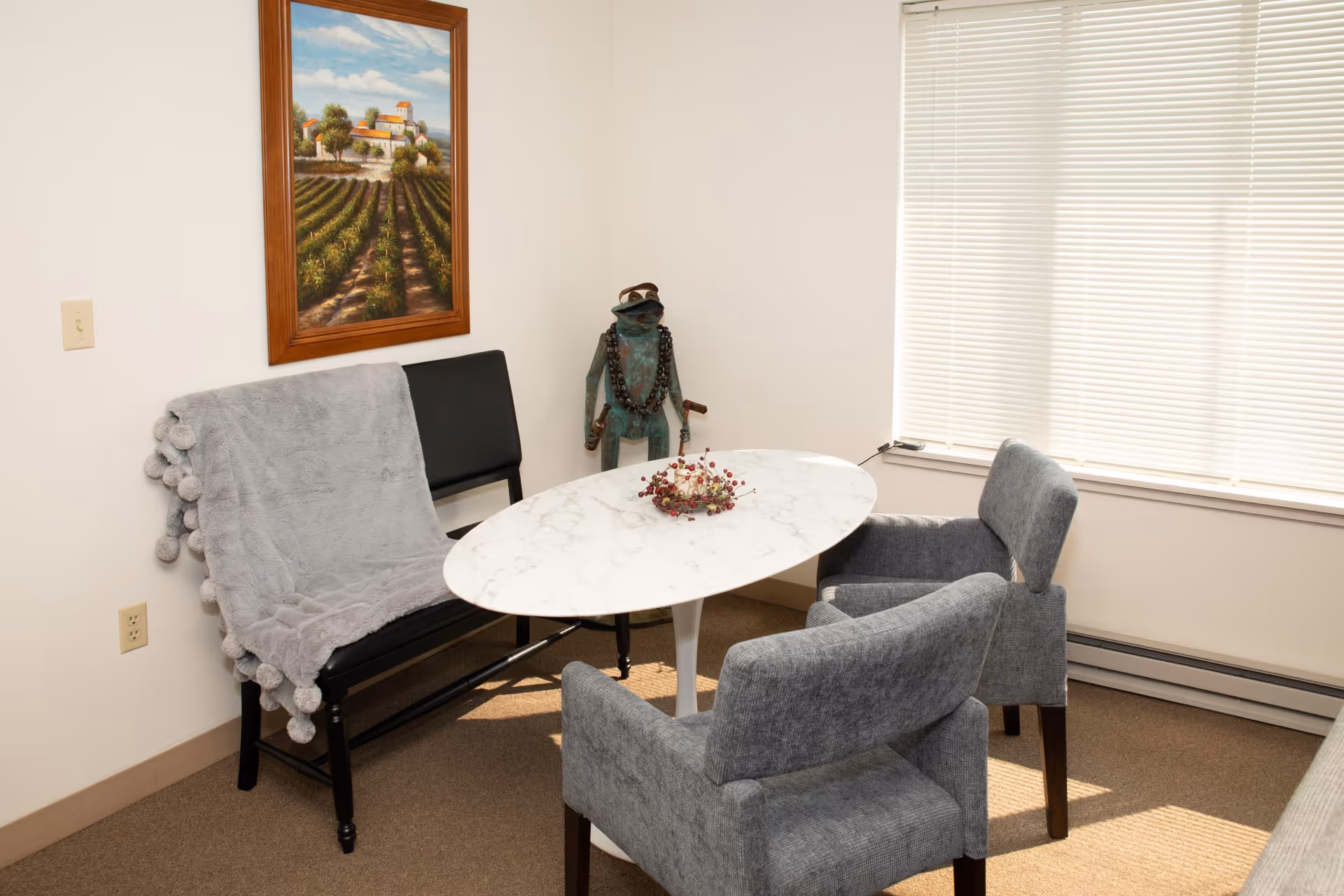 A small seating area with a white oval marble table surrounded by two gray upholstered chairs and a black bench with a gray throw blanket. A decorative frog statue wearing sunglasses and a beaded necklace stands in the corner. A framed painting of a vineyard hangs on the wall above the bench, and a window with closed blinds lets in natural light.