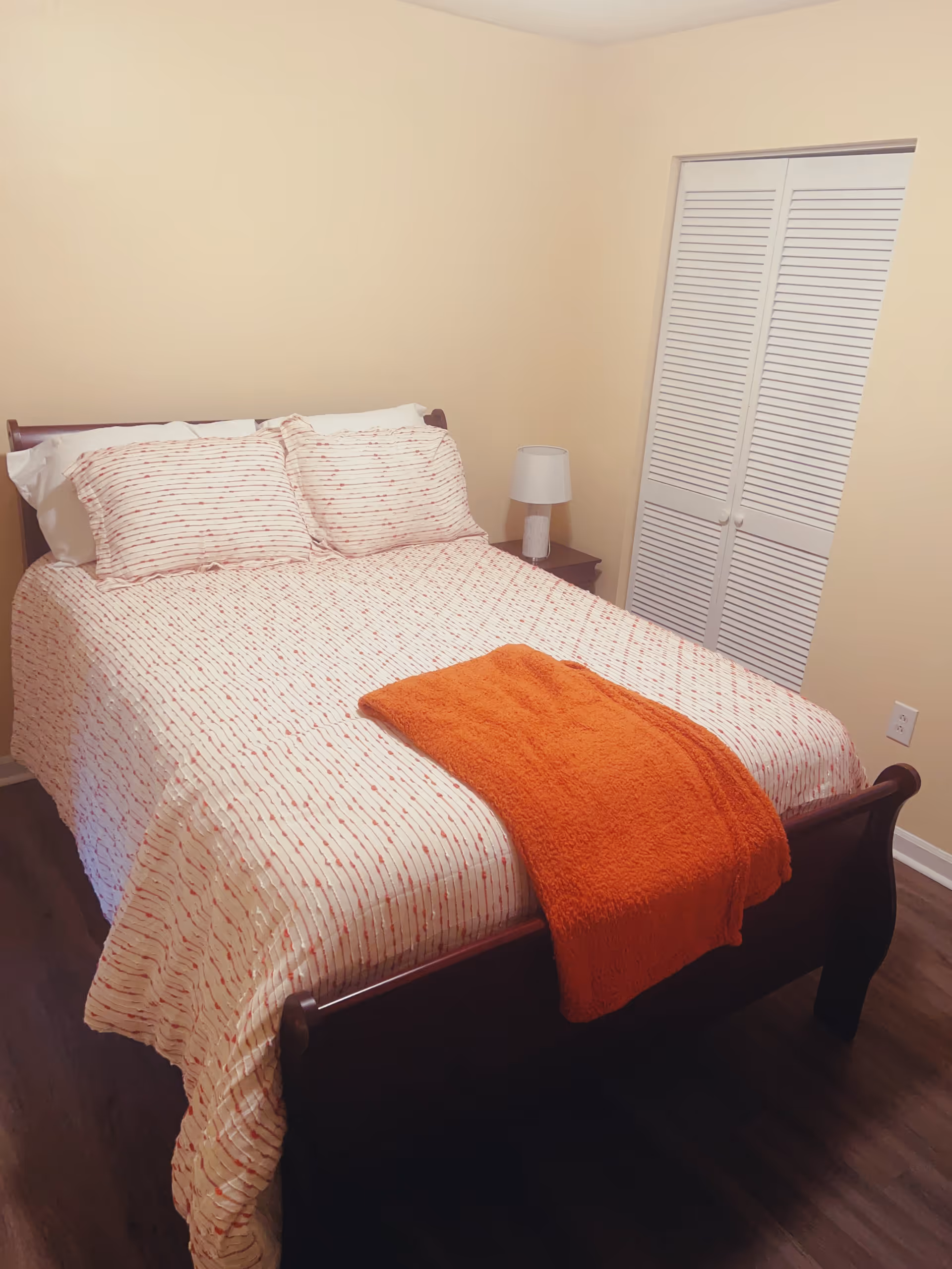 A bedroom with a wooden bed frame, a bed made with white and red patterned bedding, two pillows, and an orange blanket folded at the foot of the bed. There is a small wooden nightstand with a white lamp next to the bed, and a white louvered closet door on the beige wall.