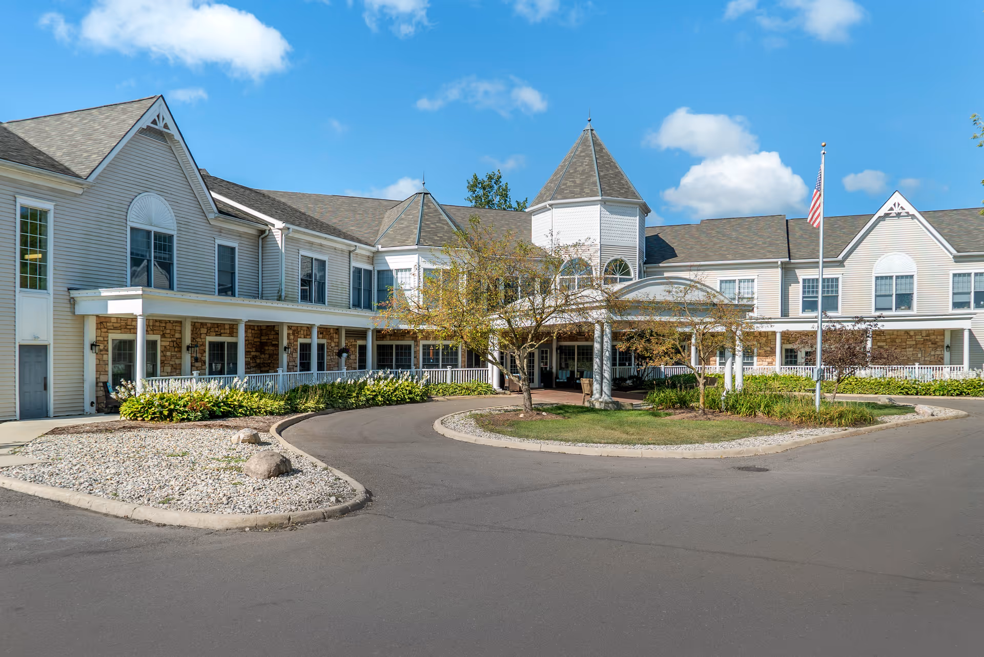 Exterior front view of a senior living facility building with beige siding, stone accents, and a covered entrance with columns. There is a circular driveway with landscaping including trees, bushes, and rocks. An American flag is visible on a flagpole near the entrance under a blue sky with some clouds.