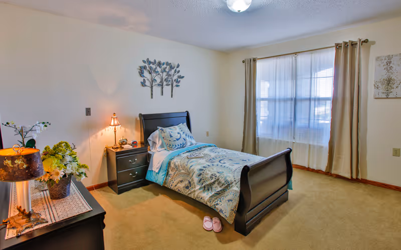 Well-lit bedroom with a single sleigh bed, nightstand, dresser with lamps and floral decor, and a window with curtains.