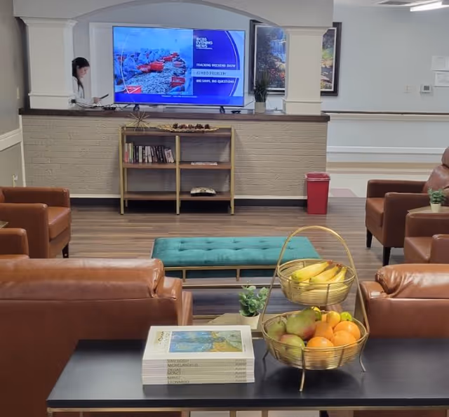 A cozy living room area in an assisted living facility with brown leather chairs arranged around a teal cushioned bench. A flat-screen TV is mounted on a white brick wall above a small wooden bookshelf with books and decorations. In the foreground, a black table holds a stack of art books and a two-tiered basket filled with bananas, pears, and oranges. The room has wood flooring and light-colored walls with framed artwork and a red trash bin in the background.
