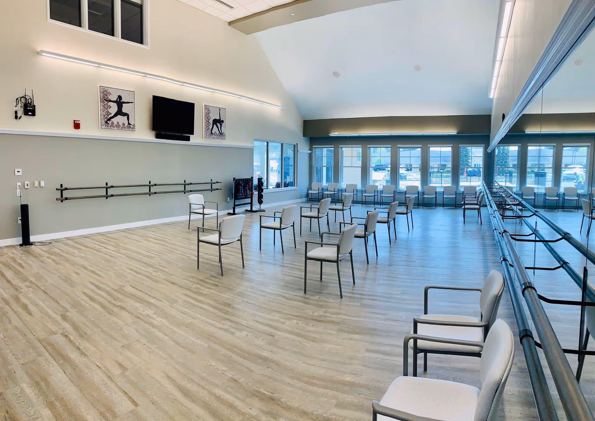 A spacious exercise or activity room with wooden flooring and multiple chairs spaced apart. The room features large windows letting in natural light, a wall-mounted TV, ballet barres along the walls, and two framed pictures of yoga poses. There is a large mirror on one side of the room reflecting the chairs and windows.