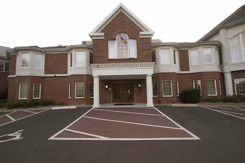 Brick two-story senior living facility entrance with columns and a paved parking area.