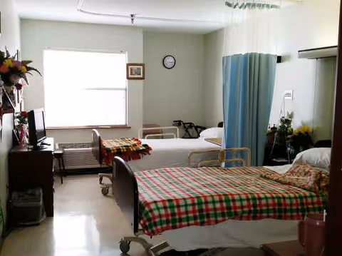 A shared patient room in a senior living facility with multiple beds and curtains.
