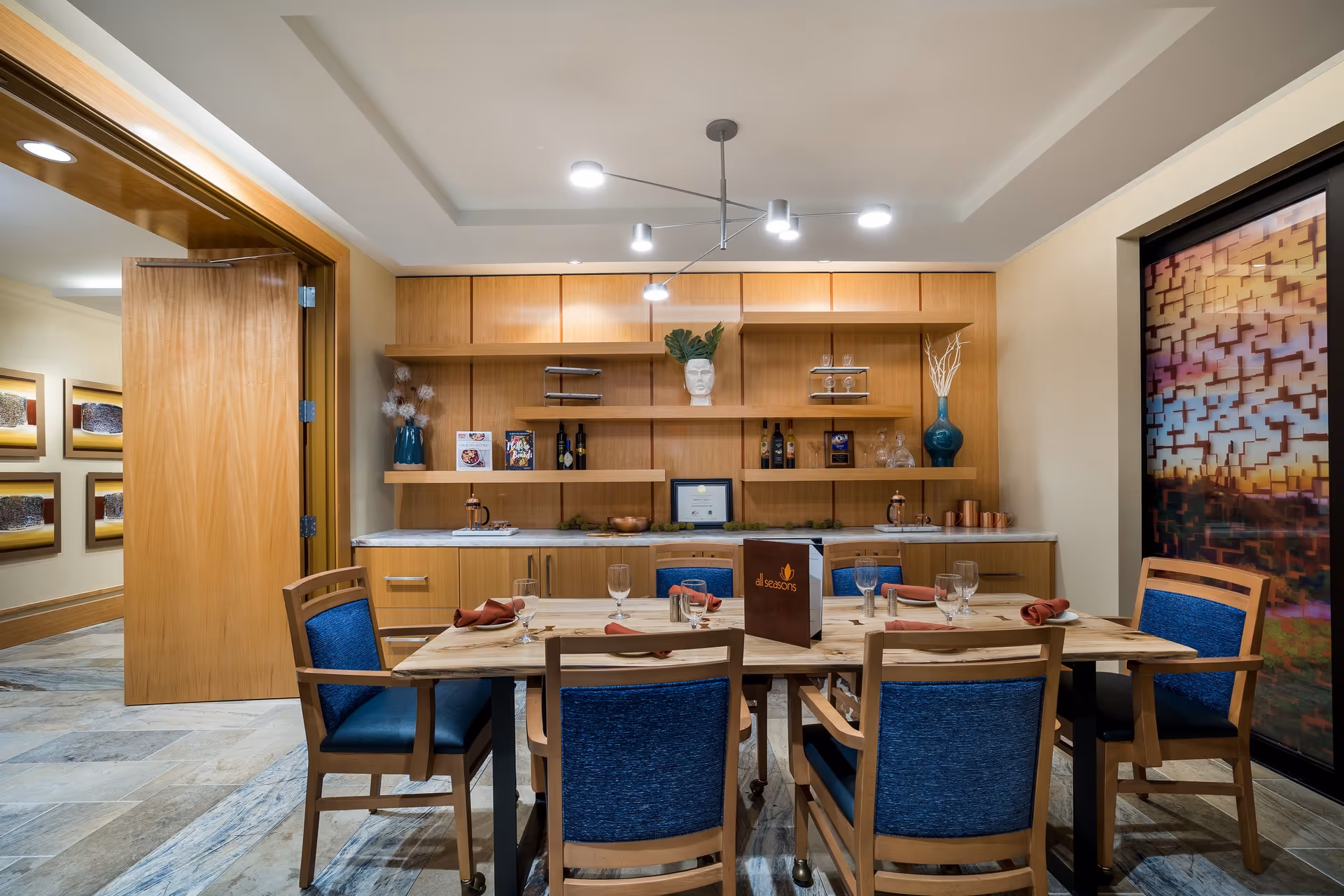 A dining area with a wooden table set for six people with blue cushioned chairs. The table has glassware, napkins, and a menu labeled 'all seasons'. Behind the table is a wooden cabinet with shelves displaying decorative items, bottles, and framed pictures. To the right, there is a large window with a colorful patterned design. The floor is tiled, and the ceiling has modern lighting fixtures.