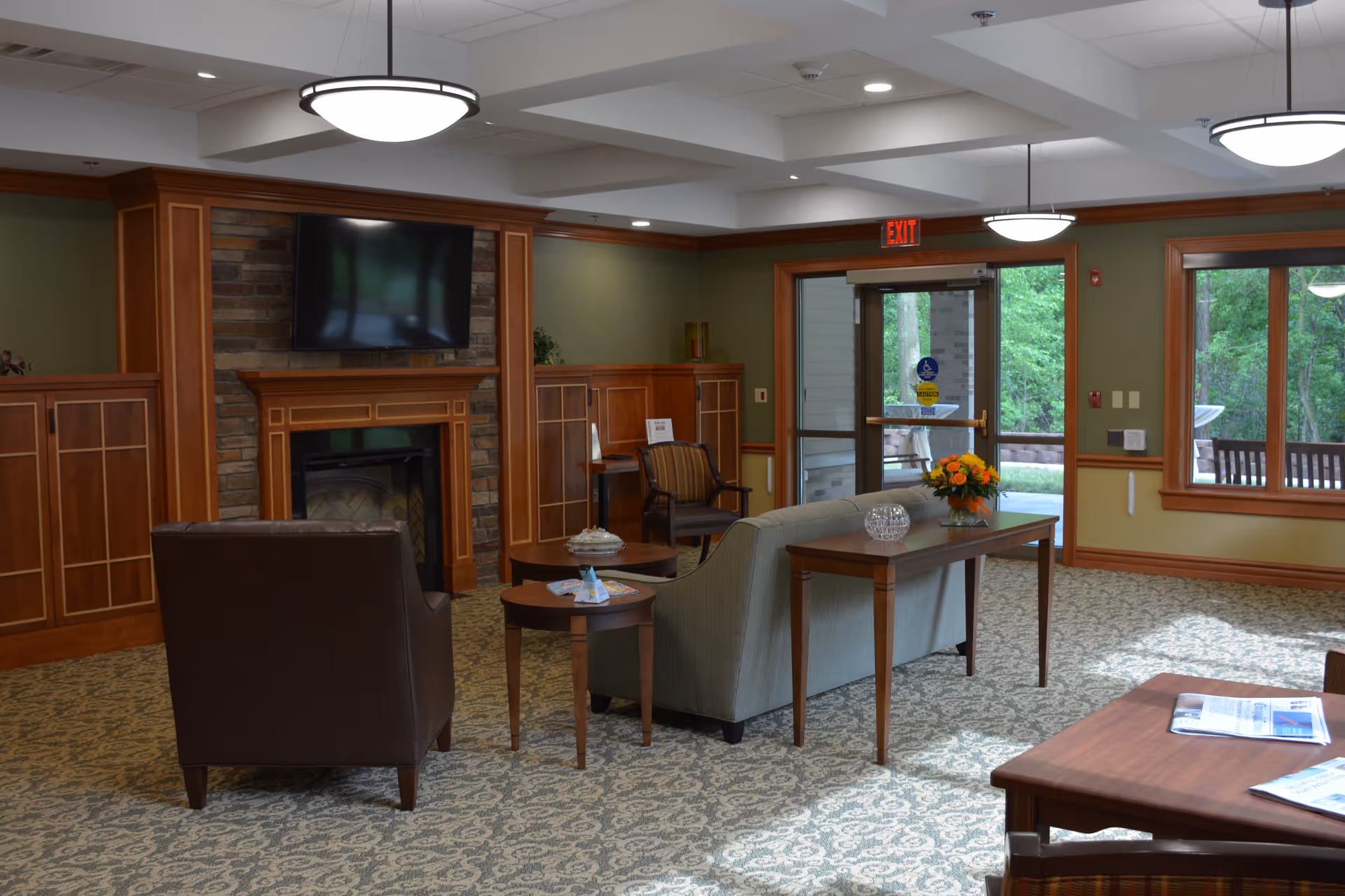 A cozy living room area in a senior living facility with a fireplace and a mounted flat-screen TV above it. The room features wooden cabinetry and trim, green walls, a patterned carpet, and several pieces of furniture including a brown leather armchair, a gray sofa, a wooden coffee table with magazines, and a side table with a flower arrangement. Large windows and a glass door provide a view of greenery outside.