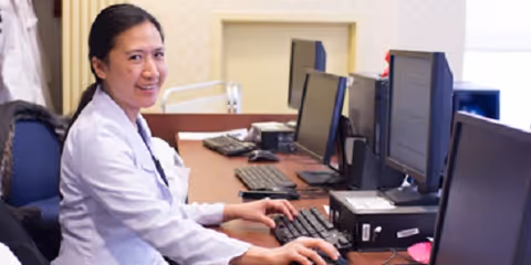 A woman in a white lab coat sitting at a desk with multiple computer monitors, smiling at the camera in an office setting.