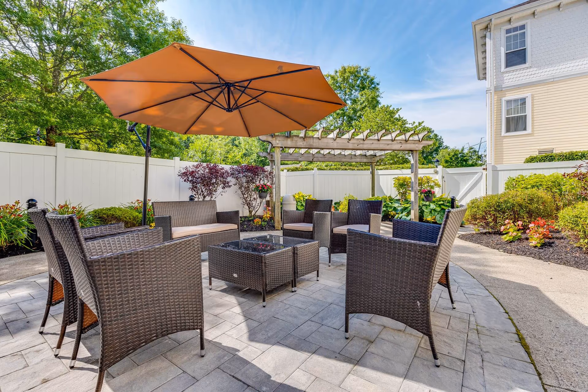 Outdoor patio area with a large orange umbrella, surrounded by several dark wicker chairs and a matching coffee table. The patio is paved with stone tiles and enclosed by a white fence. There are green shrubs, plants, and a wooden pergola in the background, with a light yellow building visible on the right side.