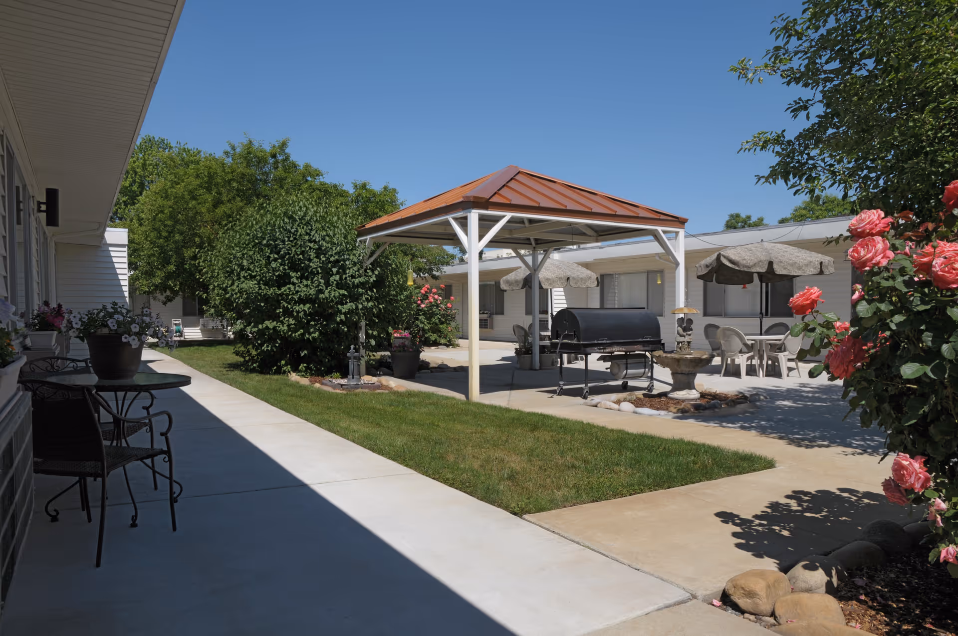 Sunny courtyard with a central gazebo, grill, patio seating, and flowering shrubs between single-story buildings.