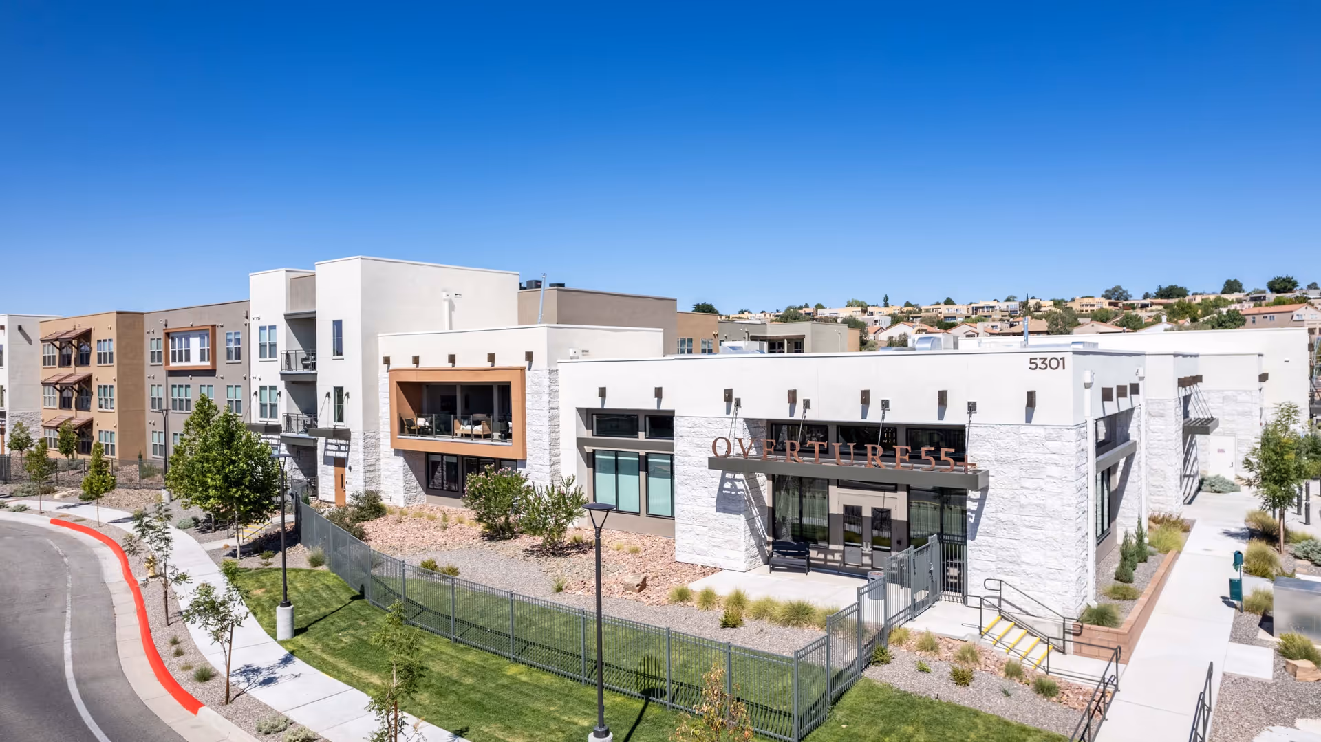Exterior view of a modern senior living facility named Overture 55, featuring a multi-story building with balconies, large windows, and landscaped surroundings under a clear blue sky.