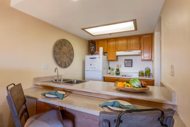 Small kitchen with a breakfast bar, two bar stools, double sink, refrigerator and wooden cabinets, decorated with a large wall clock and a bowl of fruit.