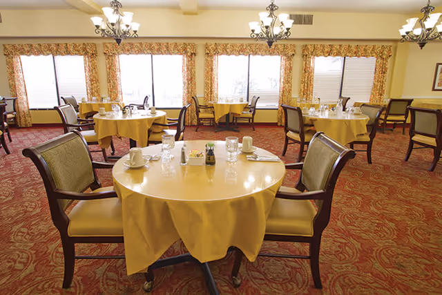 Dining room with round tables set for service, upholstered chairs, chandeliers, and patterned curtains and carpet.