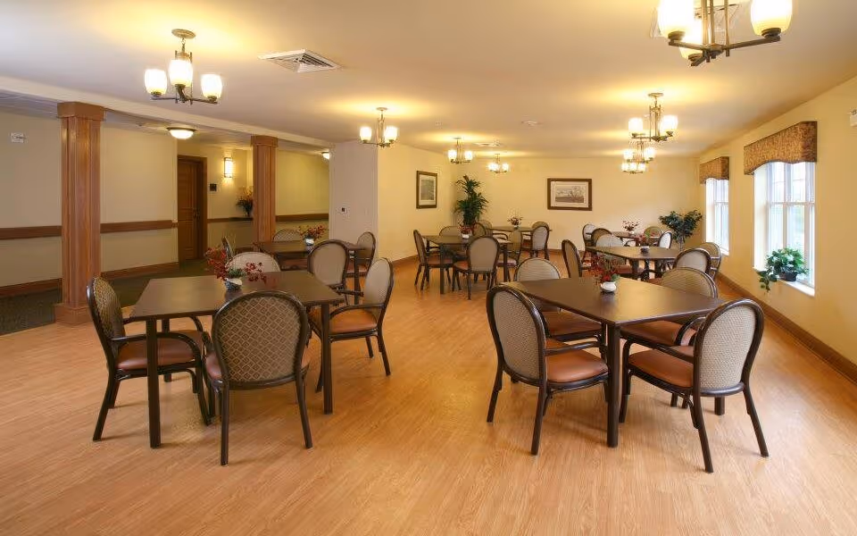A spacious dining room with multiple square tables, each surrounded by four cushioned chairs. The room has wooden flooring, beige walls, and several ceiling light fixtures. There are windows on the right side with valances and some potted plants placed near the windows and on the tables. The room appears clean and well-lit.
