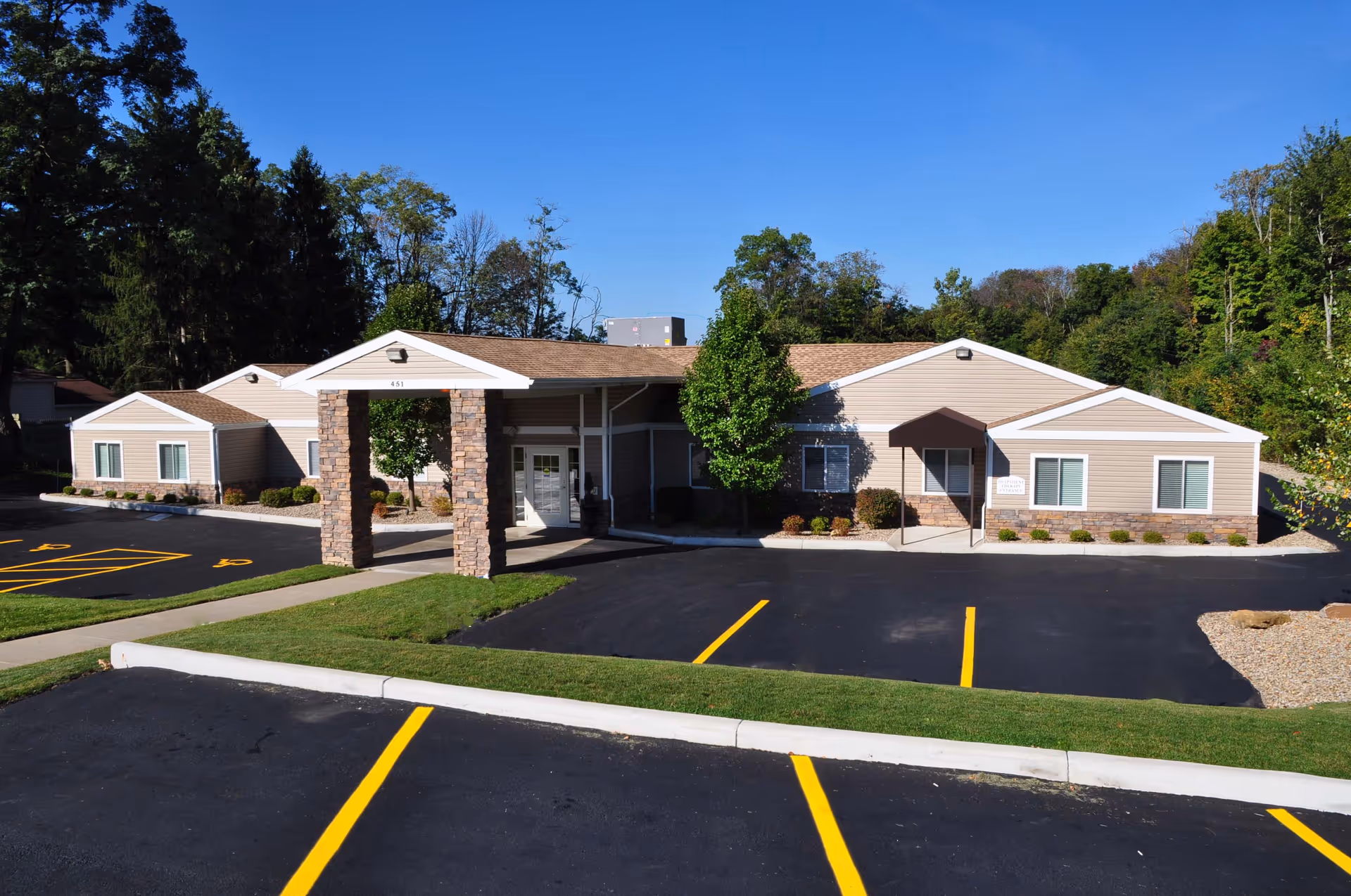 Exterior view of a single-story building with beige siding and a brown shingled roof, featuring a covered entrance supported by stone pillars. The building is surrounded by a paved parking lot with yellow parking lines and some green grass areas. Trees and a clear blue sky are visible in the background.