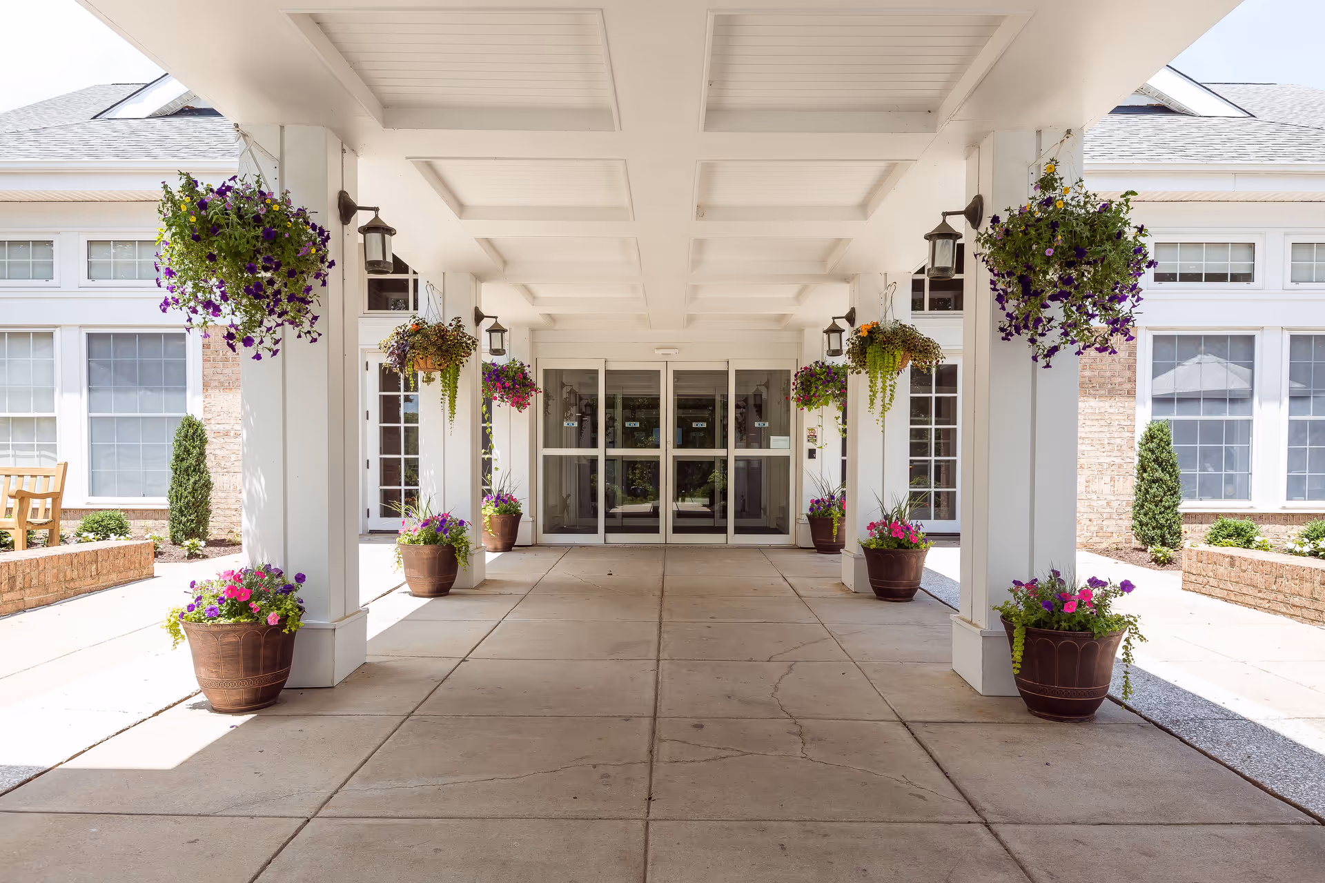 Covered entrance walkway to a building with white pillars adorned with hanging flower baskets and large flower pots at the base. The building has large windows and glass double doors at the end of the walkway.
