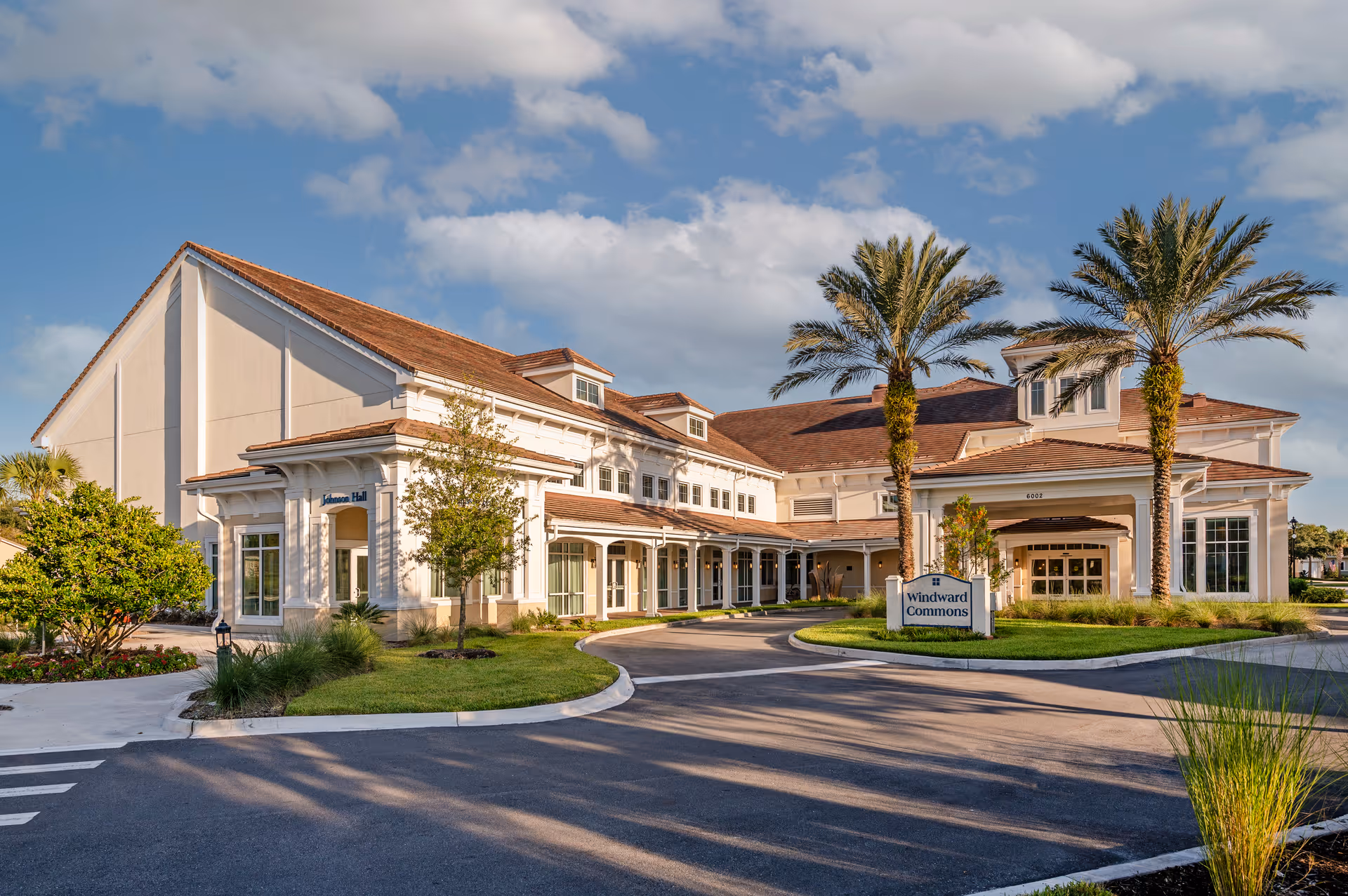Exterior front entrance of a two-story senior living community with a covered porte-cochère, palm trees, and a circular driveway.