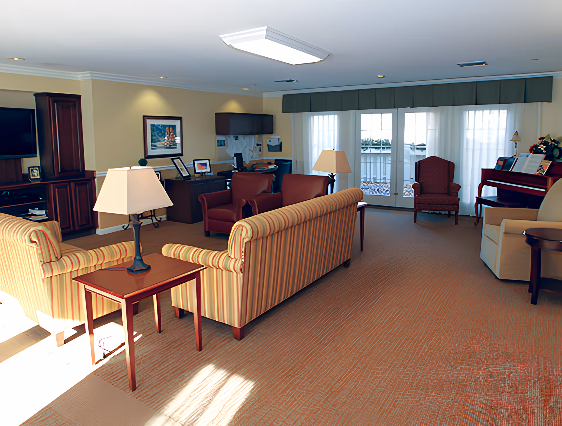 Spacious furnished common living room with striped sofas, armchairs, lamps, a TV cabinet, a piano, and large windows/doors letting in light.