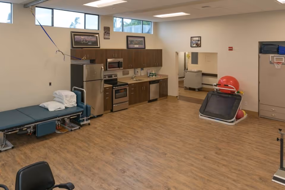Spacious rehab/activity room featuring a small kitchen area, treatment table, exercise balls and other therapy equipment on wood-look flooring.