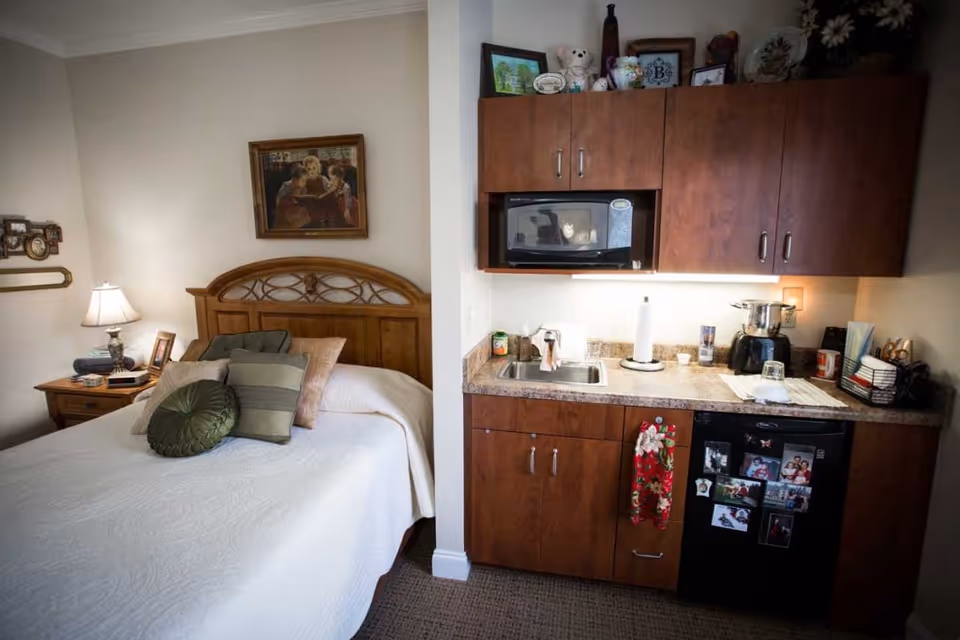 Studio-style assisted living interior showing a bed with pillows on the left and a kitchenette with cabinets, microwave, sink and mini-fridge on the right.