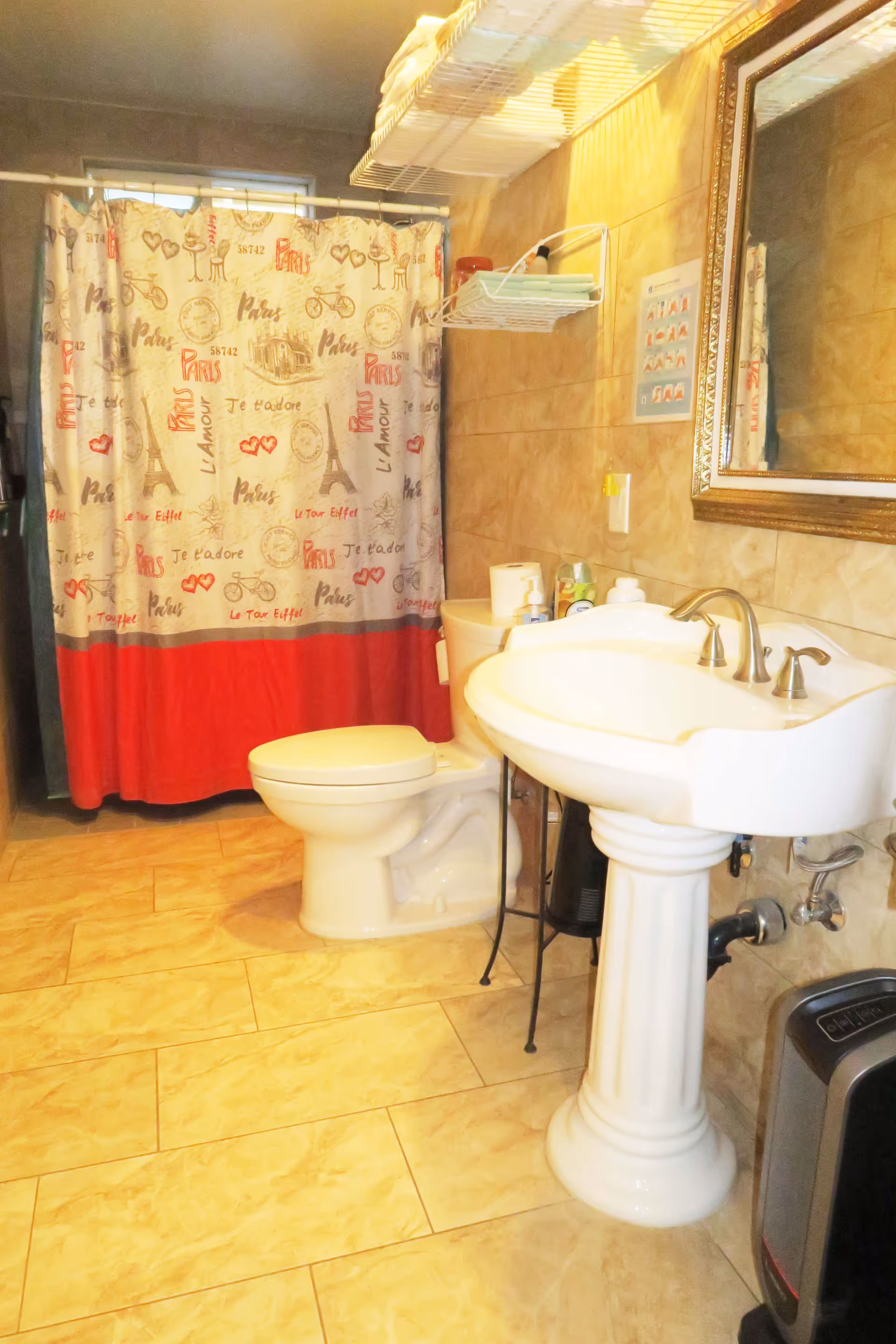A bathroom with beige tiled walls and floor, featuring a white pedestal sink with silver faucets, a toilet, and a shower with a curtain decorated with Paris-themed designs including the Eiffel Tower and French words. There is a mirror above the sink and a white wire shelf mounted on the wall holding towels and toiletries.