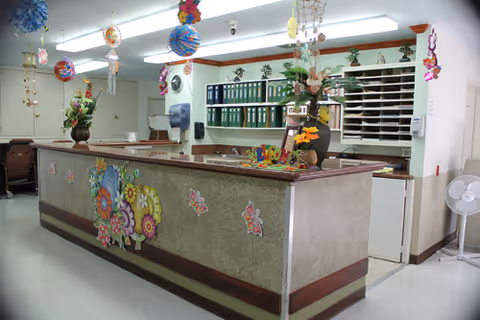 Reception desk area decorated with colorful floral and butterfly stickers, hanging paper decorations from the ceiling, and flower arrangements on the counter. Behind the desk are shelves with green binders and a window with horizontal blinds. A fan is placed on the floor to the right.