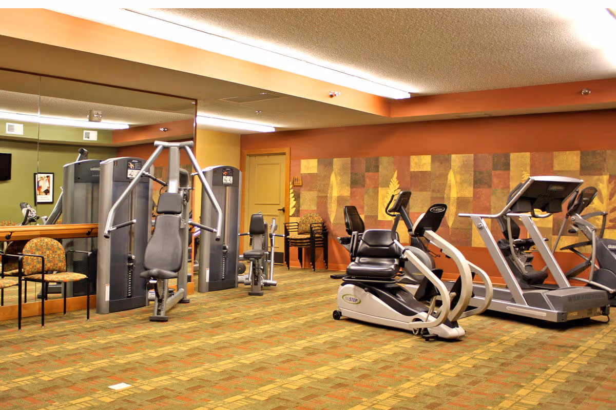 A fitness room in a senior living facility with various exercise equipment including a recumbent bike, treadmill, and weight machines. The room has a patterned carpet, a large mirror on one wall, and a decorative wall panel with leaf designs. There are also a couple of chairs along the wall.